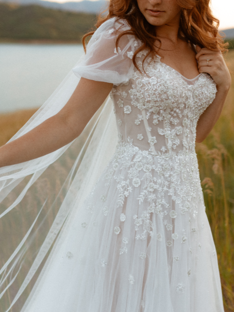 Woman in a white wedding dress with floral embellishments and a sheer cape, outdoors near a lake.