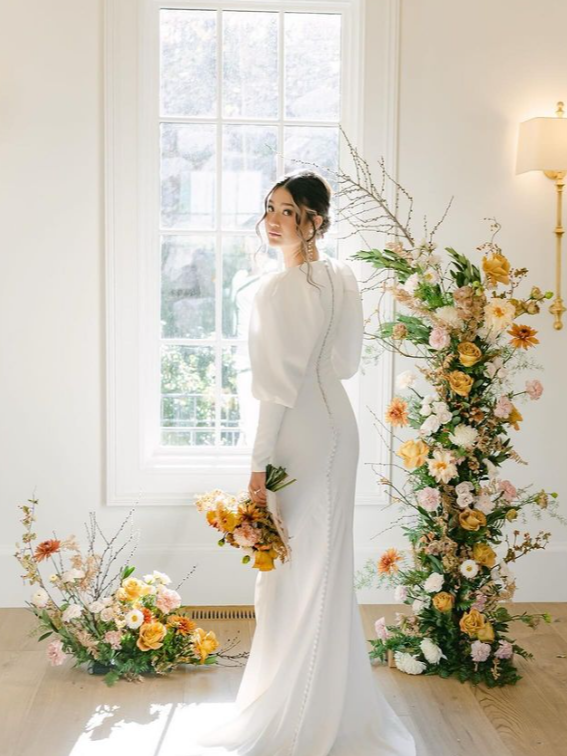 Woman in white wedding dress with floral arrangement by a window.