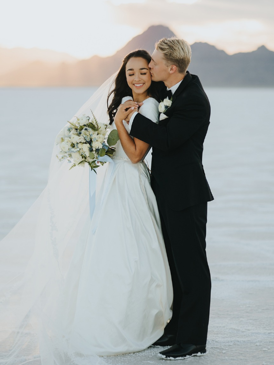 Bride and groom embrace, he kisses her cheek. They stand on a salt flat with mountains in the background.
