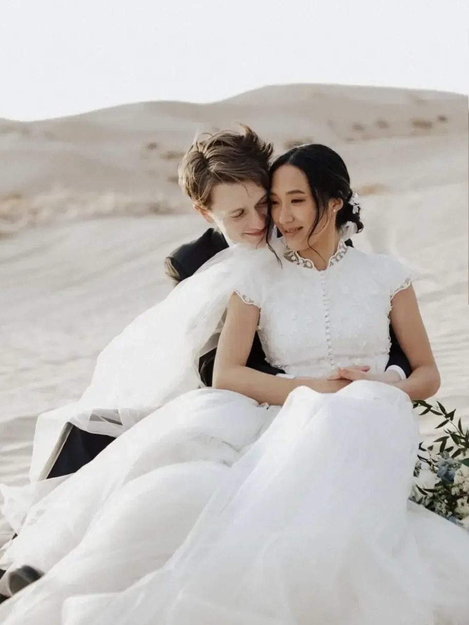 Couple in wedding attire embrace on sand dune, soft lighting.