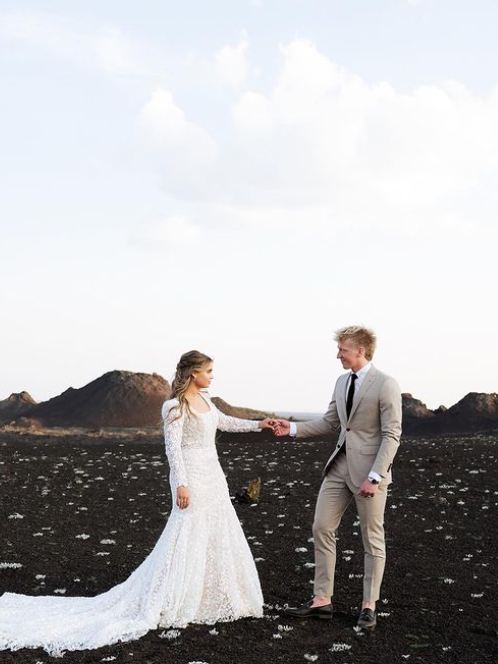 Bride and groom holding hands, standing on black volcanic landscape, blue sky.