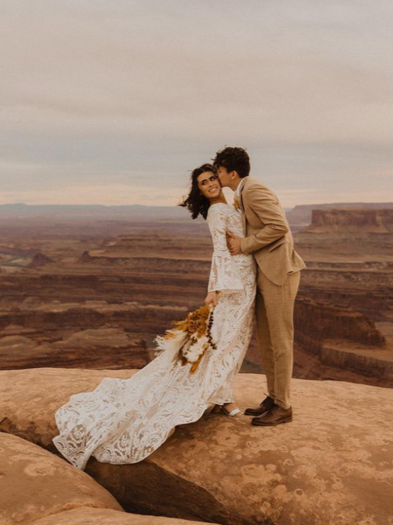 Couple kissing, bride in lace dress holding bouquet, groom in tan suit, on cliff overlooking canyon.