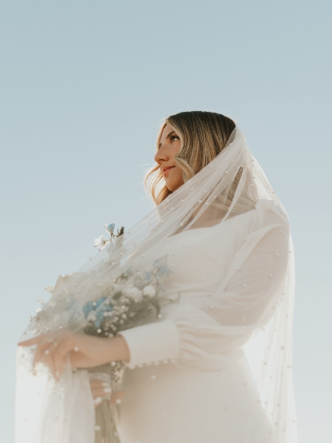 Bride in a white gown and veil, holding flowers, against a clear blue sky.