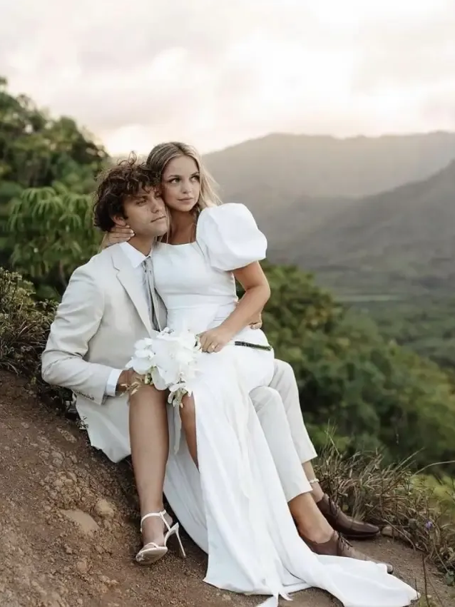 Couple in wedding attire sitting on a hillside, overlooking a valley at sunset.