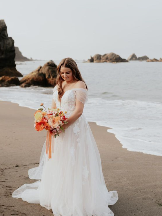Woman in wedding dress on beach, holding bouquet, looking down. Ocean waves in the background.