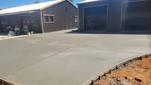 Concrete driveway in front of a dark wooden shed and large garage bays, with dirt along the edge.