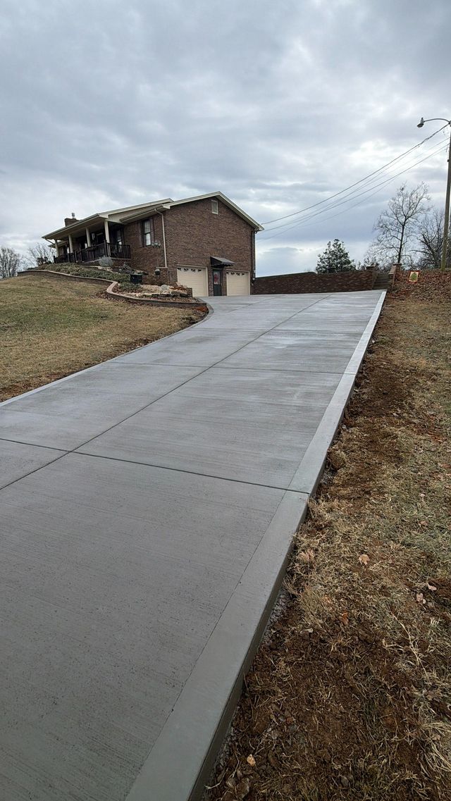 Concrete driveway leading to a brick house on a cloudy day, with grassy slopes on both sides