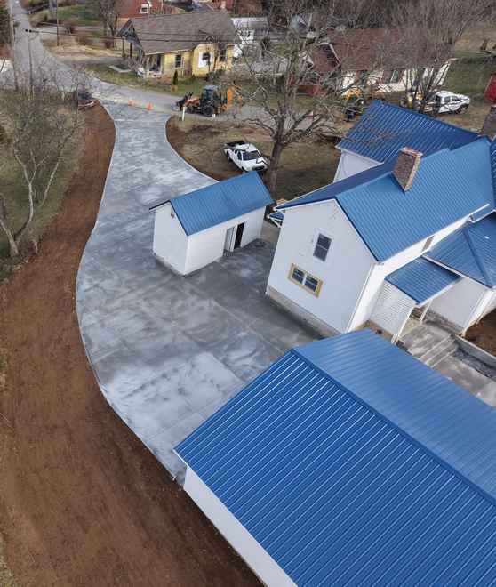 Aerial view of blue-roofed houses and a driveway beside a wooded yard under construction.