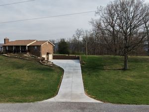 Concrete driveway leading to a brick house on a grassy hillside with leafless trees in the background