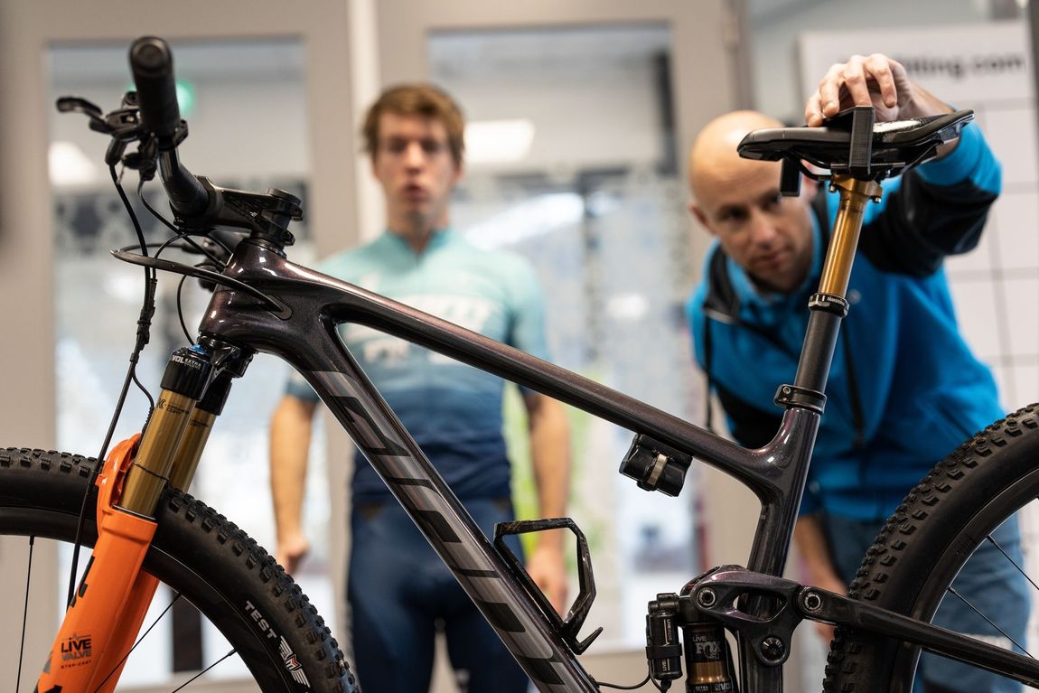 Two men are working on a bicycle in a room.
