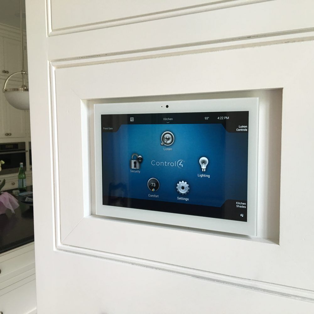 White-framed smart home control panel embedded in wall, showing Control4 interface on blue screen.
