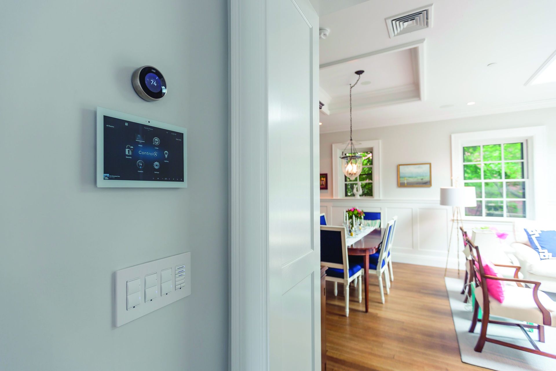 Smart home control panel, thermostat, and light switches on a white wall next to a dining room.
