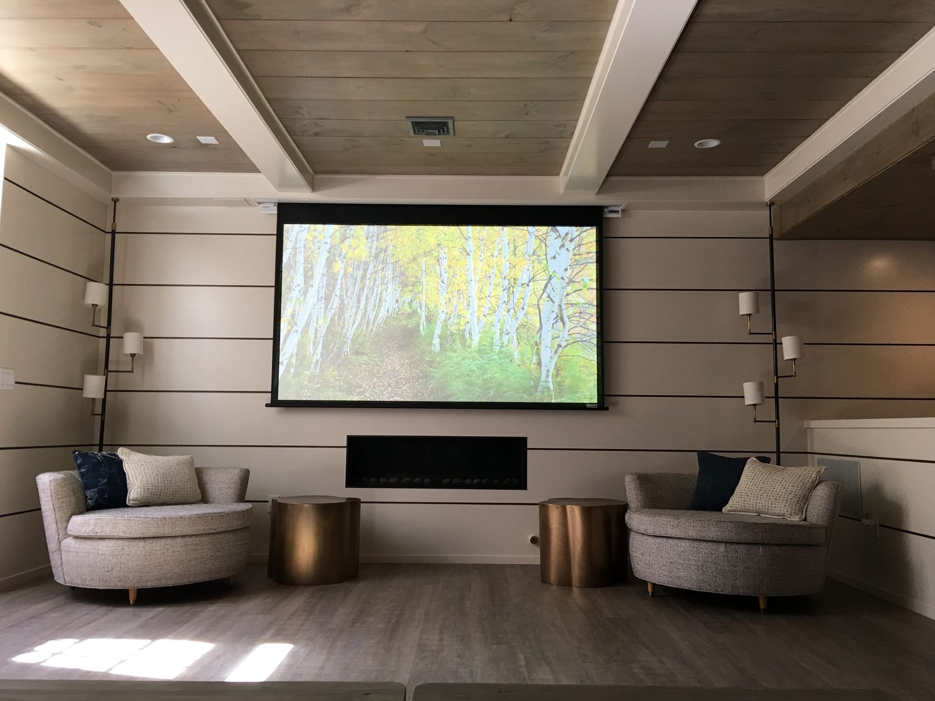 Cozy living space with two round chairs facing a screen displaying a nature scene. Fireplace below, gold side tables.