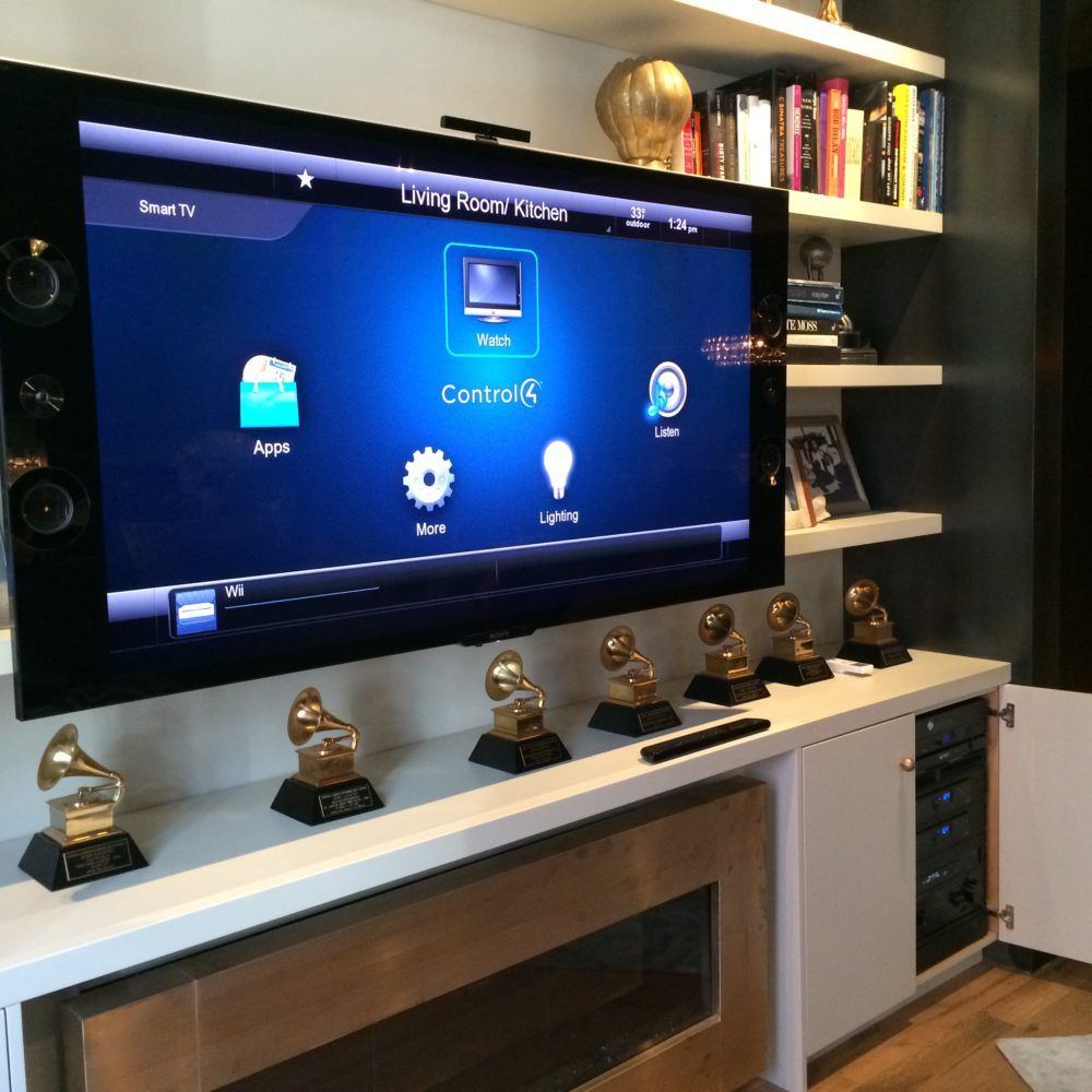 A large TV displaying a control panel, with several Grammy awards lined up below it.