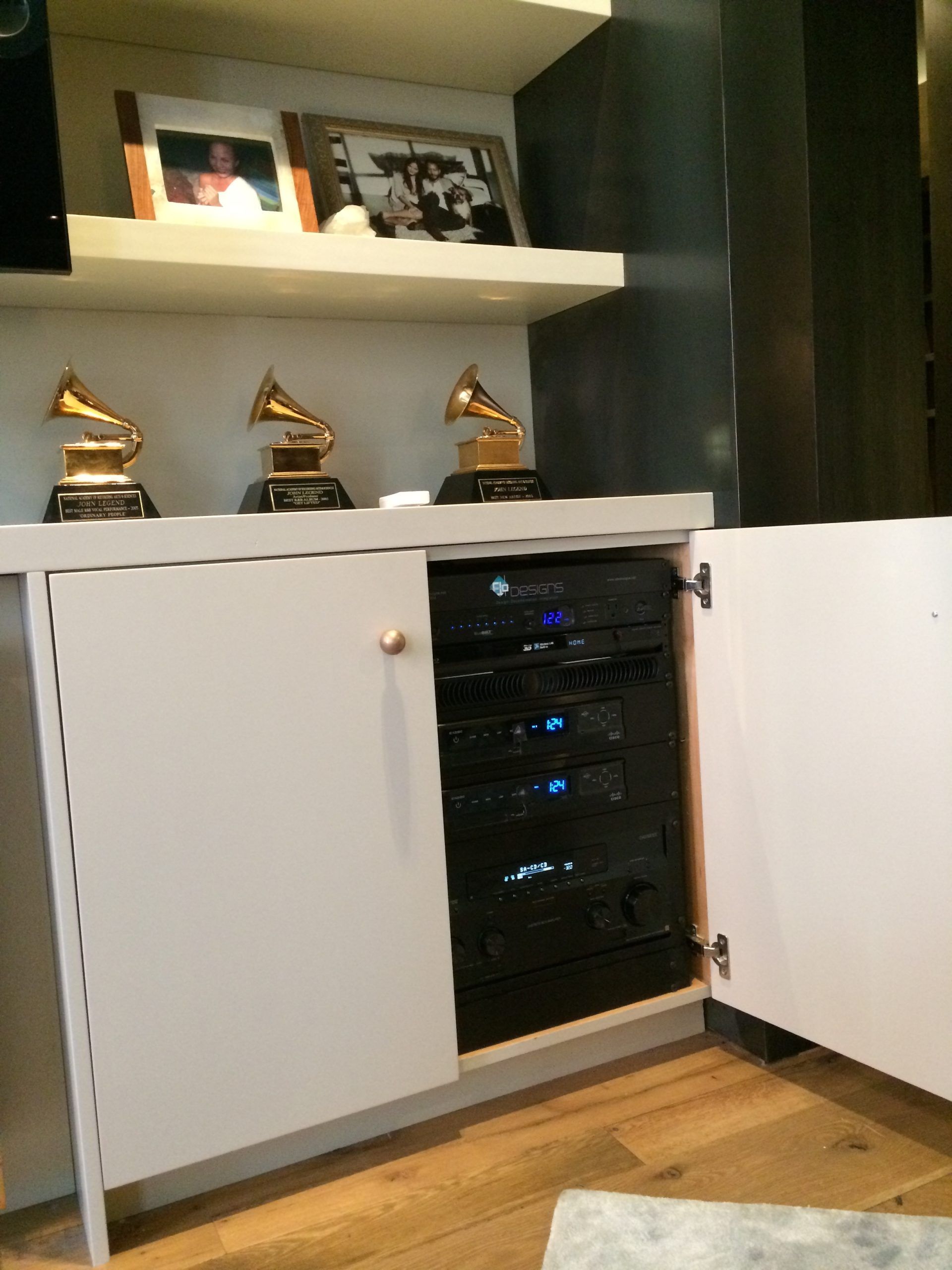 White cabinet with open door revealing black electronics. Three gold Grammy awards sit above the cabinet.