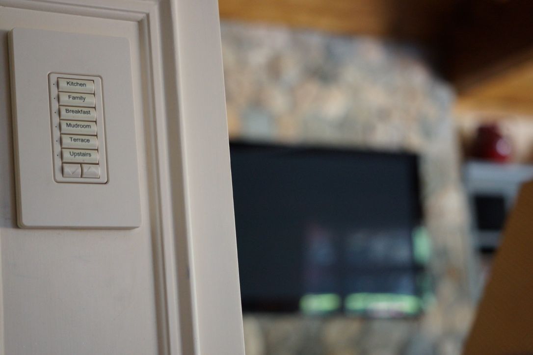 White light control panel on a wall, with a blurred TV and stone fireplace in the background.