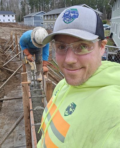 A man wearing a hat and safety glasses is taking a selfie at a construction site.