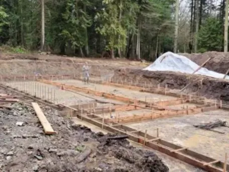Construction site: wooden forms for a concrete foundation, surrounded by dirt and trees.