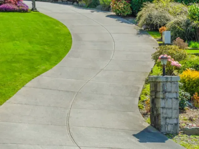 A concrete driveway leading to a house with a stone pillar.