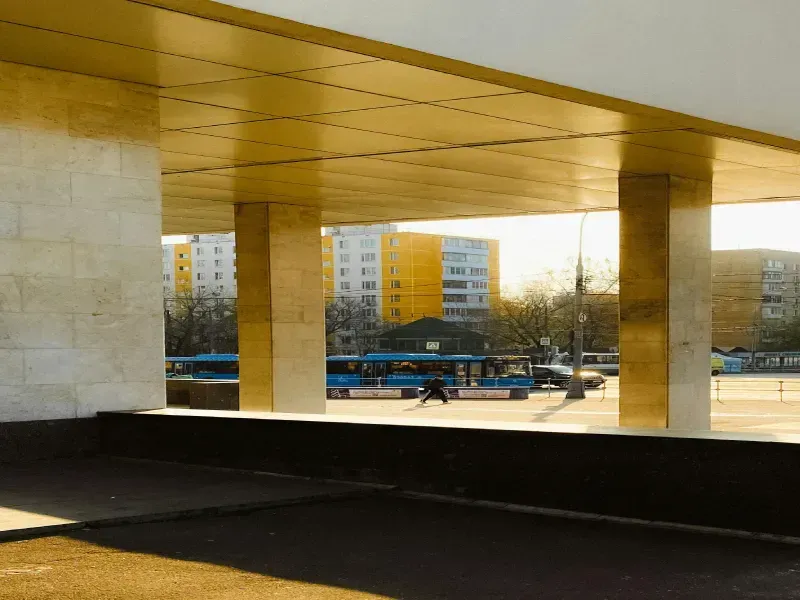 Columns and overhang frame a sunny view of buses, buildings, and a person walking in an urban setting.