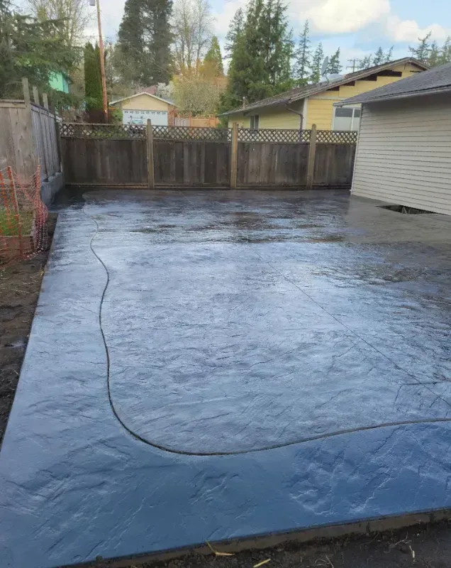 Newly poured dark gray concrete patio in a backyard, next to a wooden fence.