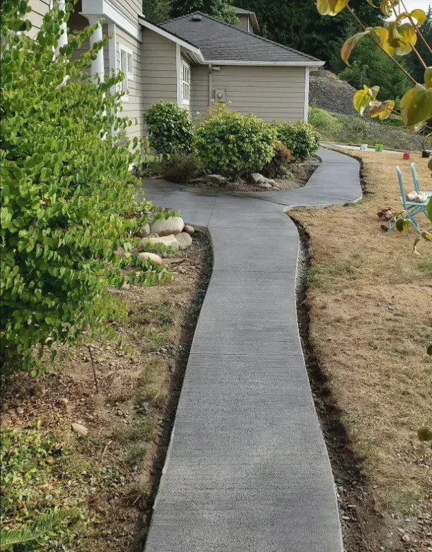 A newly poured concrete walkway curves through a front yard toward a house with tan siding and a dark shingled roof.