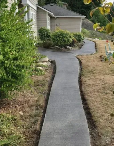 Concrete walkway winding through yard next to beige house and bushes.