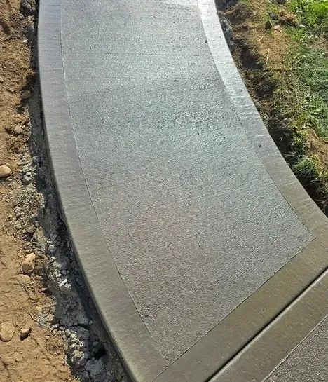 Curved concrete walkway with a rough, textured surface, next to dirt and grass.