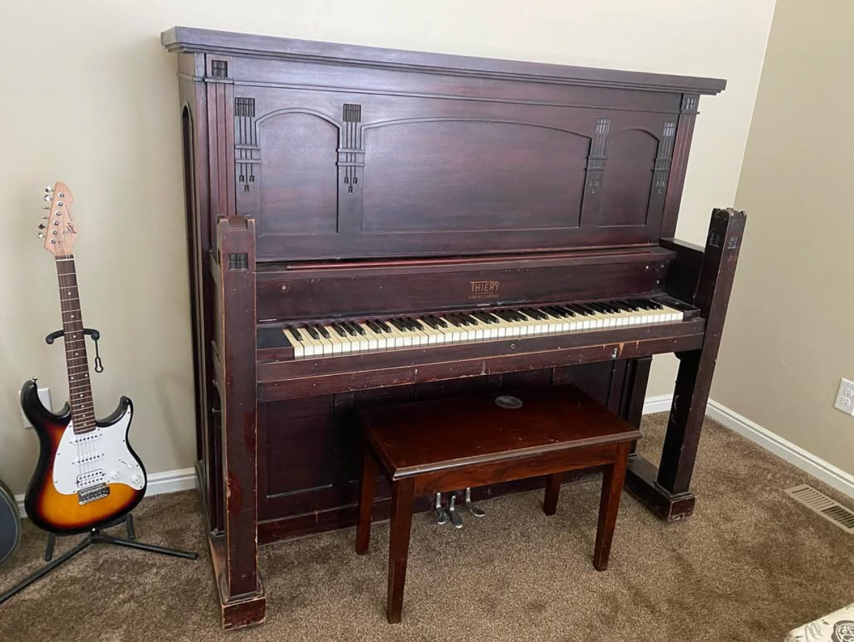 Upright piano with bench in a carpeted room, with an electric guitar leaning beside it.