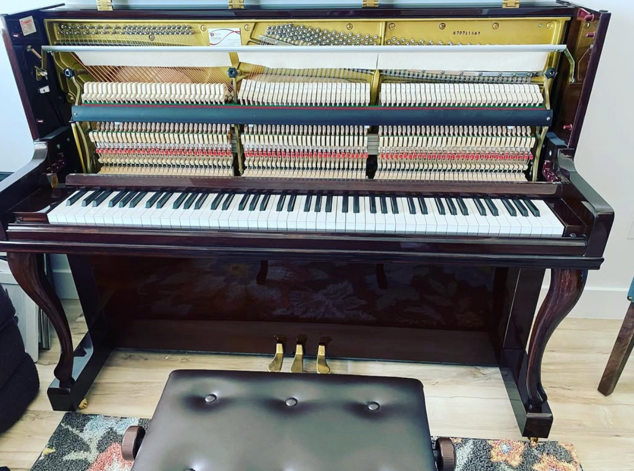 Upright piano with exposed strings and keys, sitting above a black bench in a room