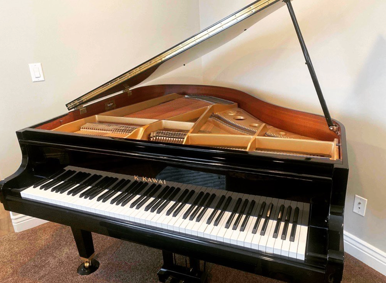 Black grand piano with raised lid, showing strings and keyboard, in a room with beige walls.