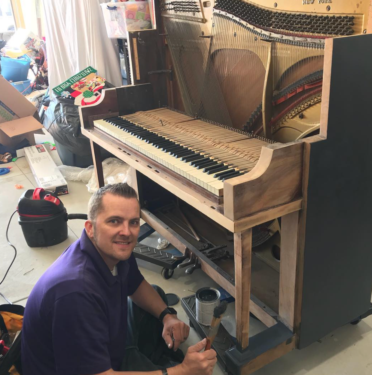 Man kneeling beside an upright piano being repaired, with exposed strings and tools in a workshop