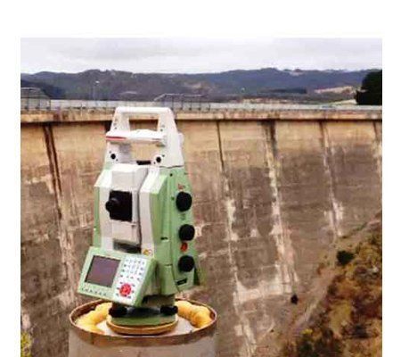 A total station is sitting on top of a barrel in front of a dam.