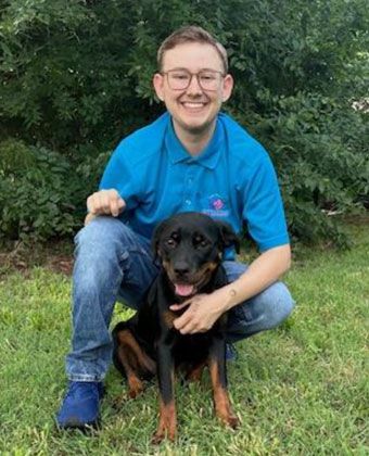 A young man is kneeling down next to a dog in the grass.