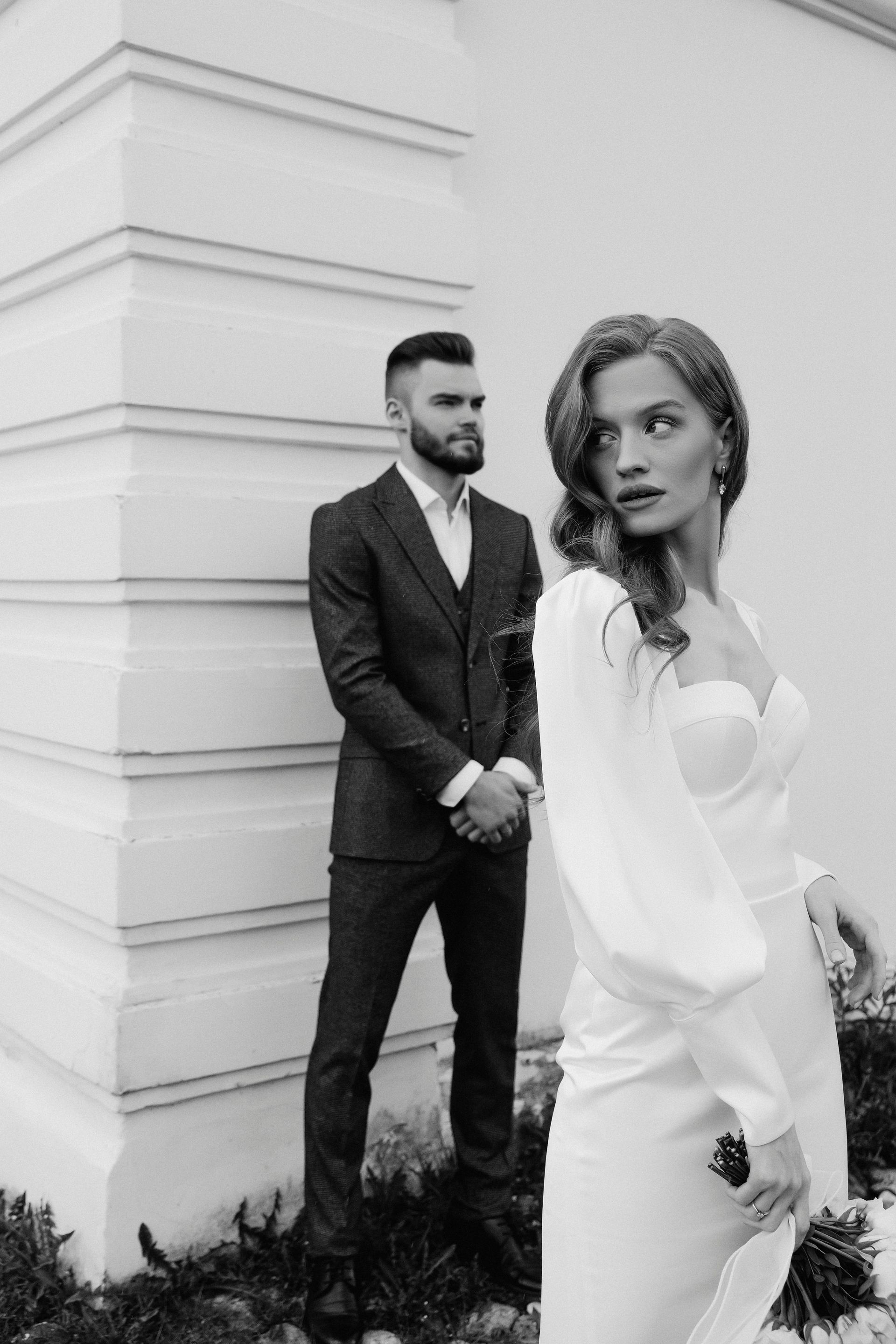 Bride in white dress looks back; groom in suit stands behind against a white building.