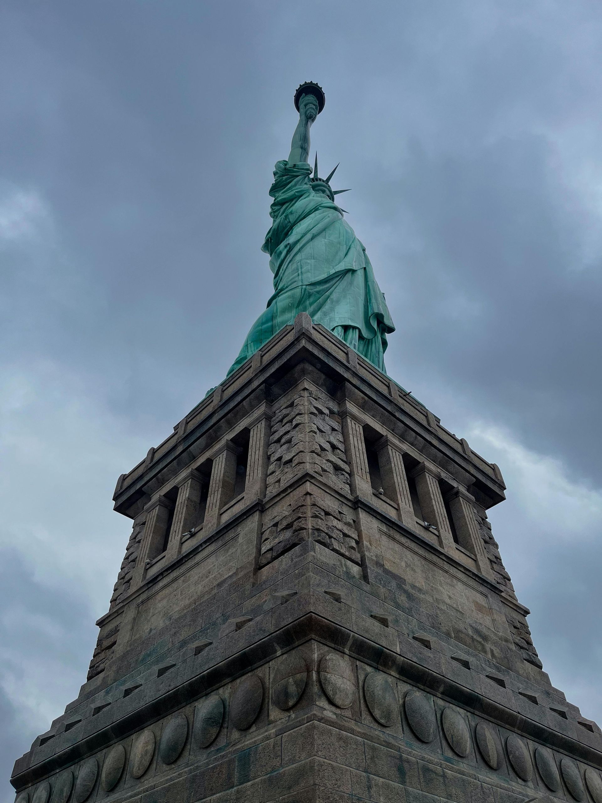 Statue of Liberty, on a stone pedestal, against a cloudy grey sky.