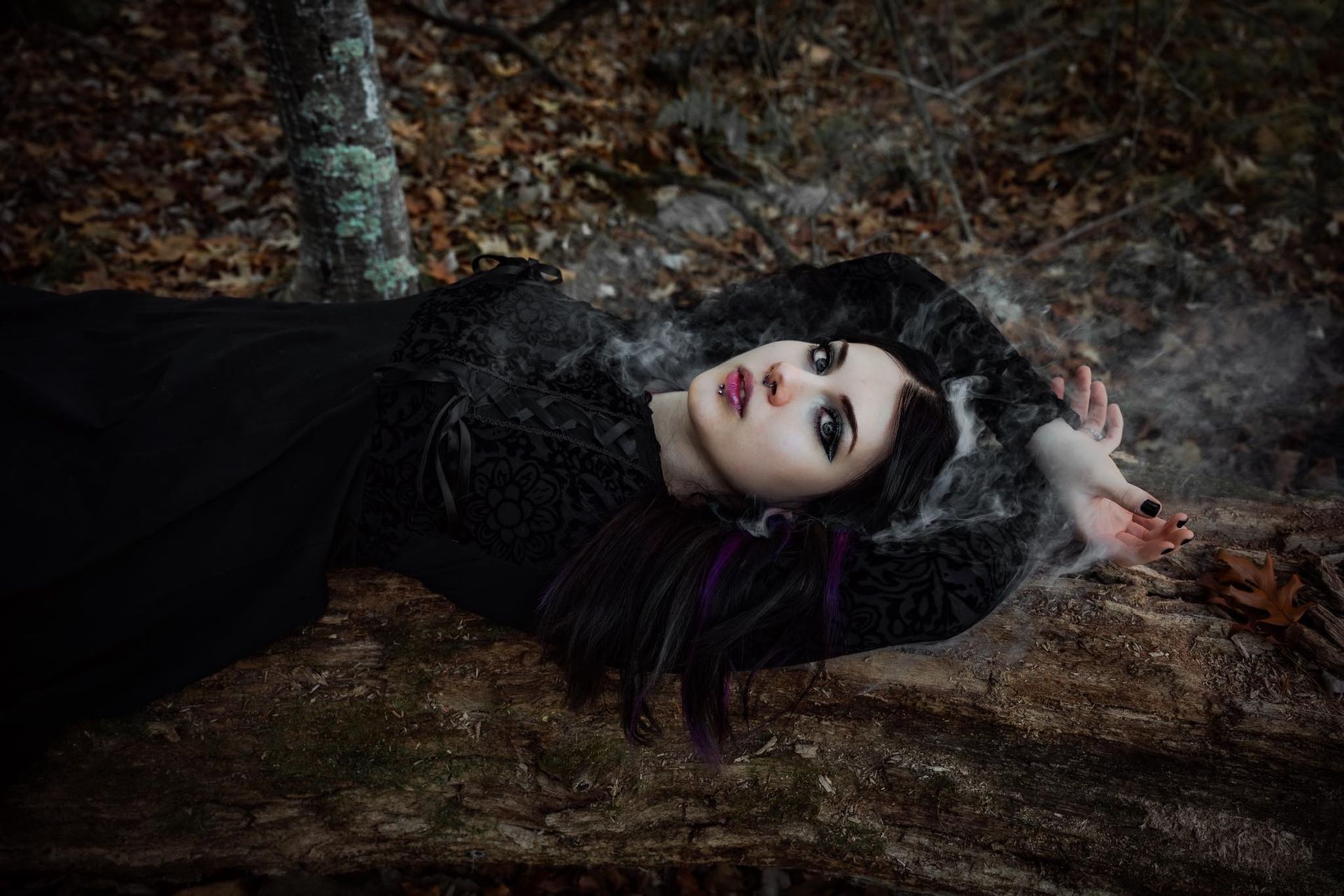 Woman in black dress and dark makeup lies on forest floor, arms outstretched, gazing upward.