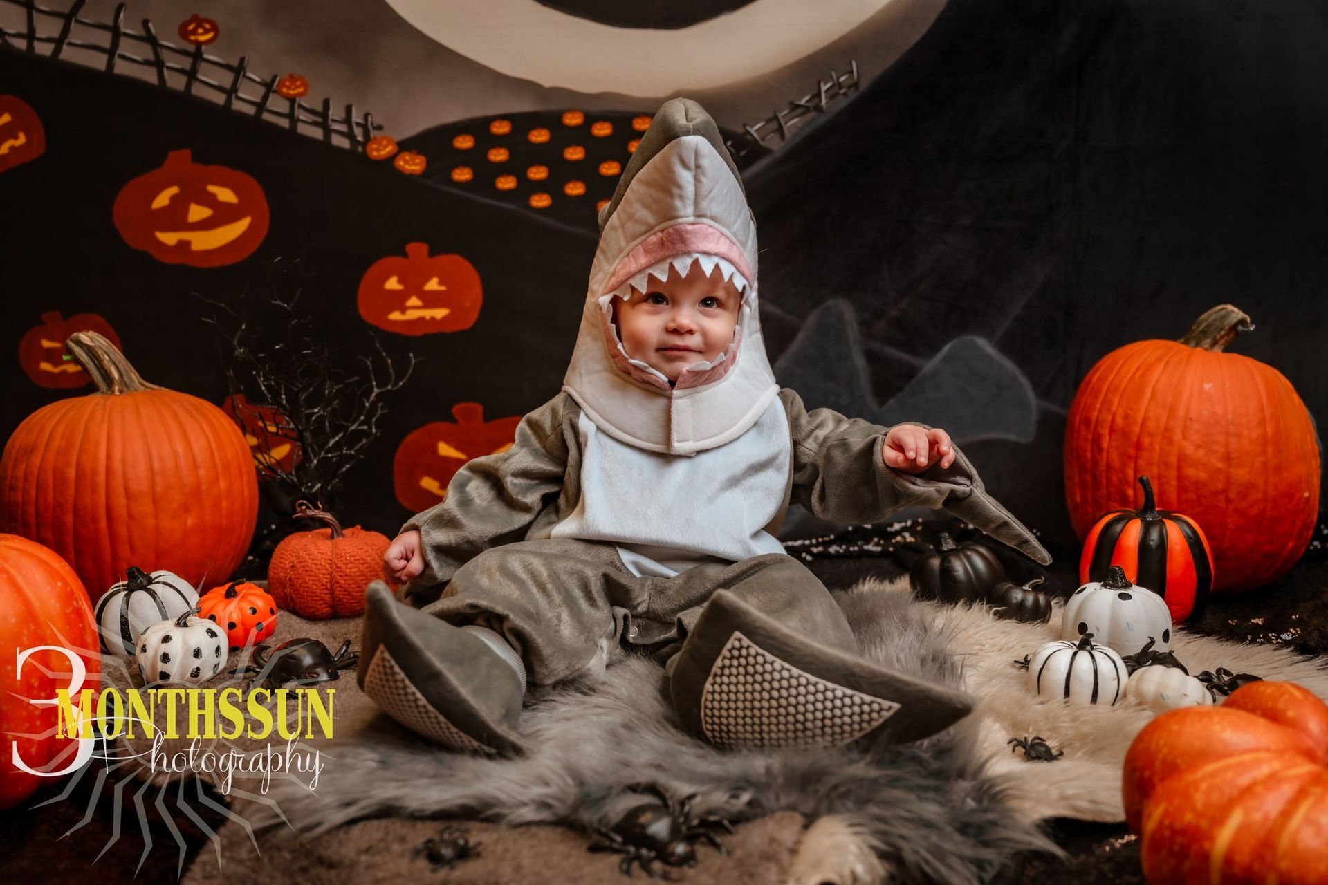 Child in shark costume sits amid pumpkins, Halloween backdrop.