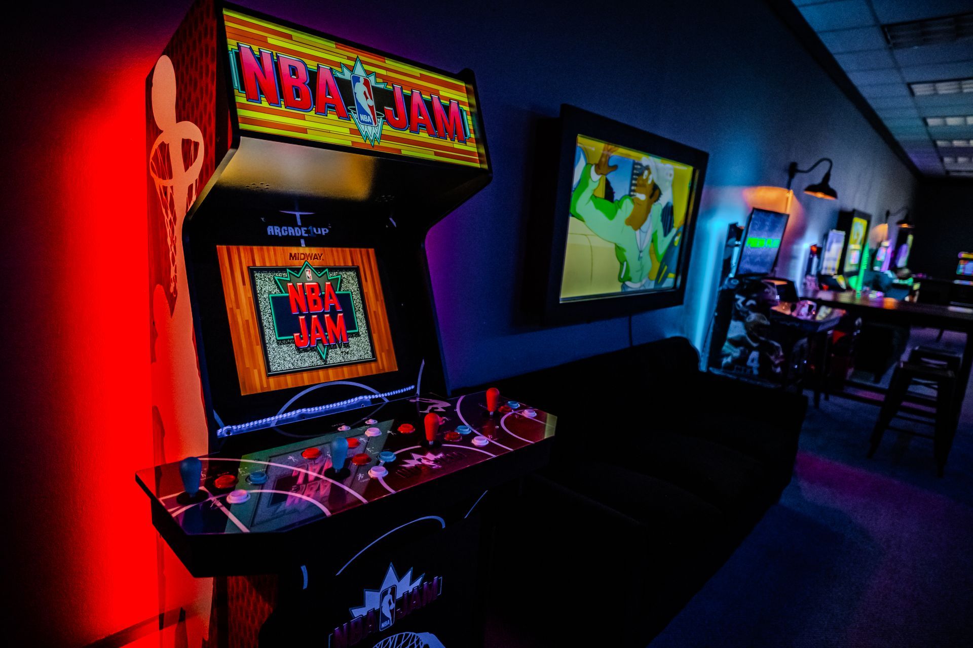 Arcade with an NBA Jam machine lit with red, blue, and purple lights. Other arcade games and a TV are in the background.