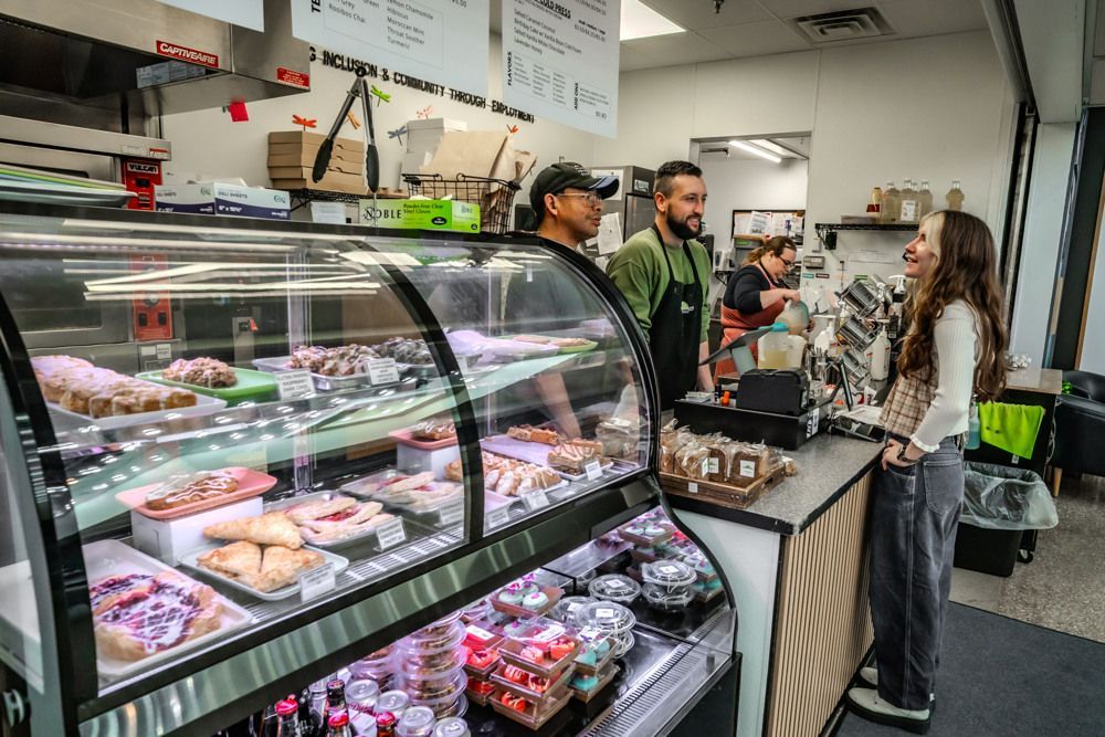 Bakery counter with pastries. Three staff members interact with a customer; the display case is full.