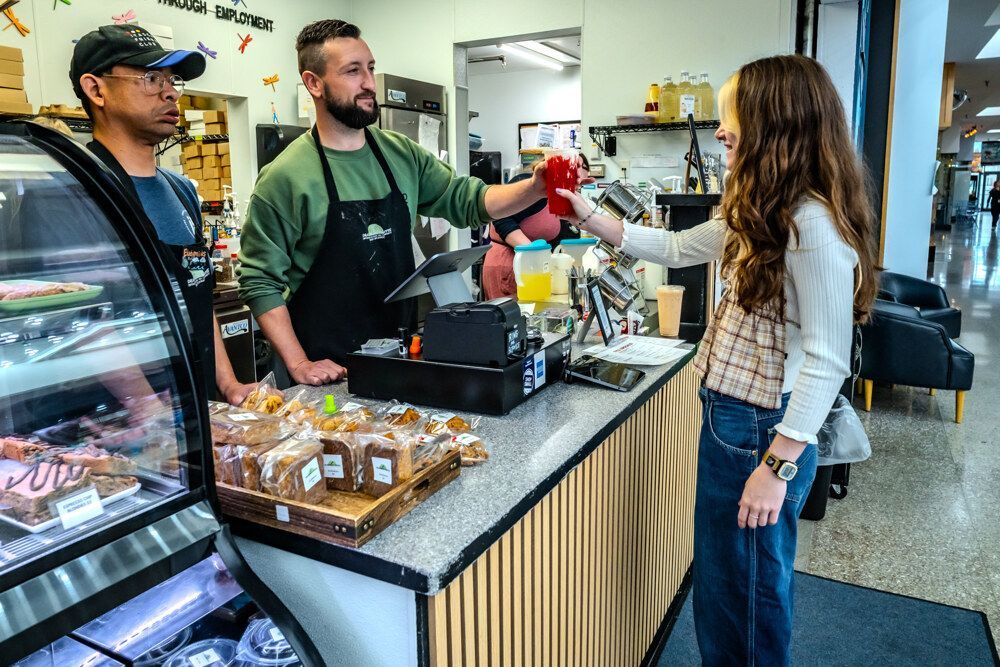 A barista hands a red drink to a customer at a cafe counter. A display case of baked goods is visible.