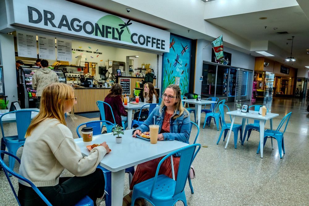 Two women seated at table at Dragonfly Coffee cafe in mall. Blue chairs, white tables.
