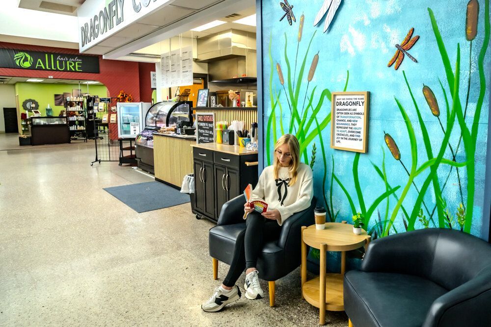 Woman reading book in a coffee shop, sitting in a black chair near a mural of reeds and dragonflies.