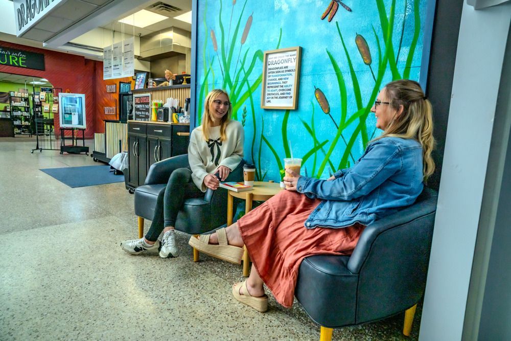 Two women chatting, seated in armchairs. One holds a beverage near a small table. Wall mural behind them.