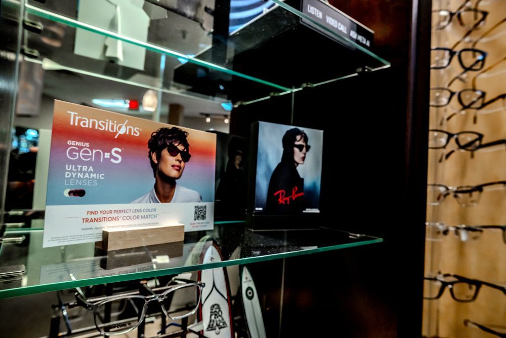 Optical shop display with Transitions Gen S and Ray-Ban ads, glasses on shelves, and reflected store interior.