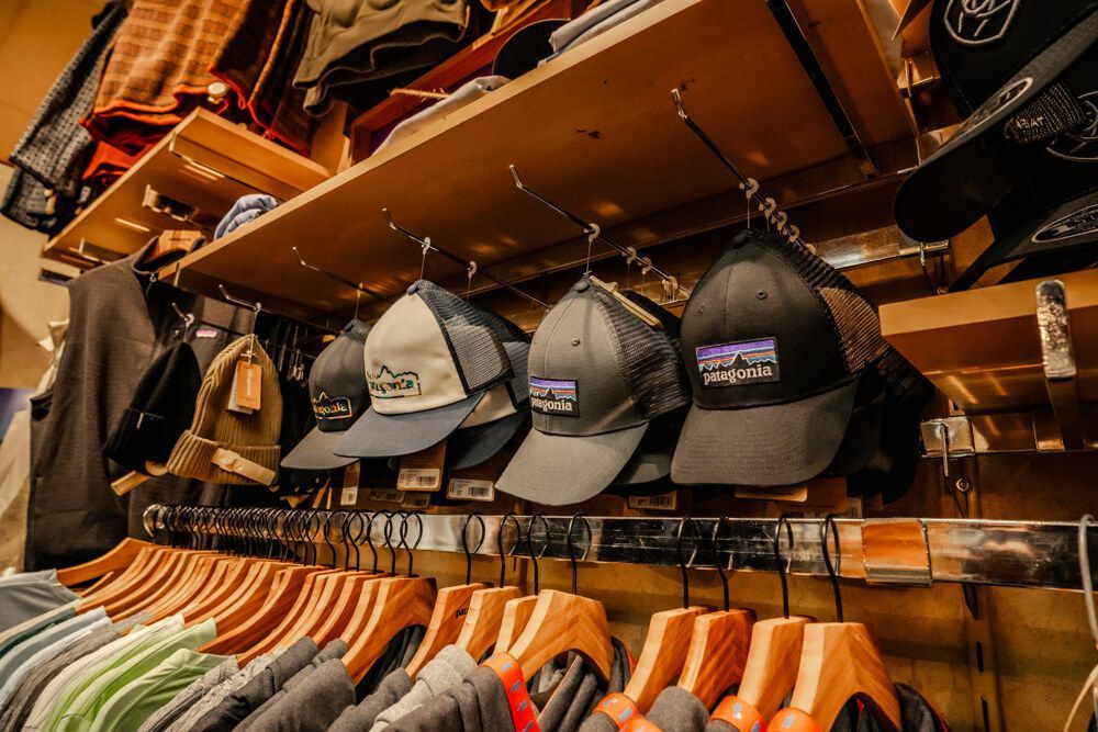 Hats and clothing on display in a store. Brown shelves and wooden hangers. Patagonia logo on the hats.