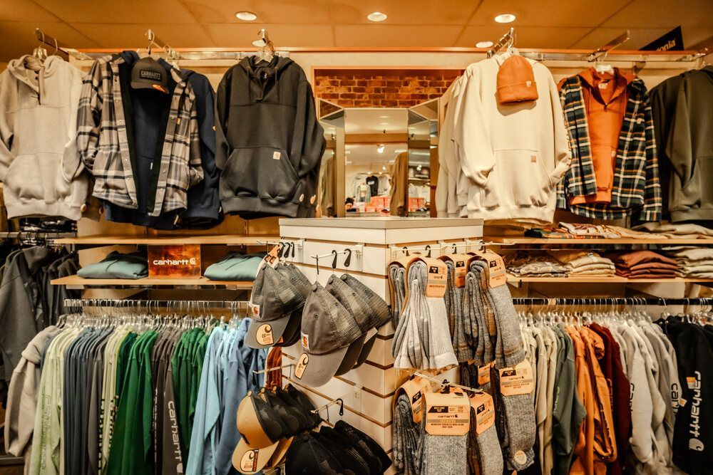Clothing store interior, with shirts, hoodies, and hats displayed on racks and shelves. Neutral colors, wood accents.