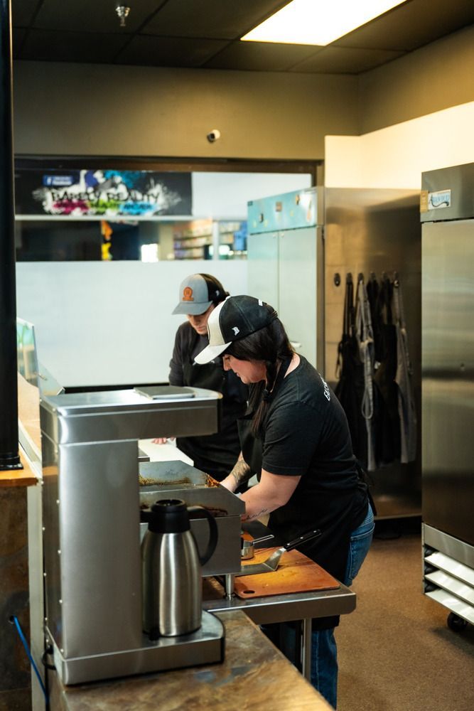 Two people preparing food in a commercial kitchen; one cutting on a cutting board, the other looking on.