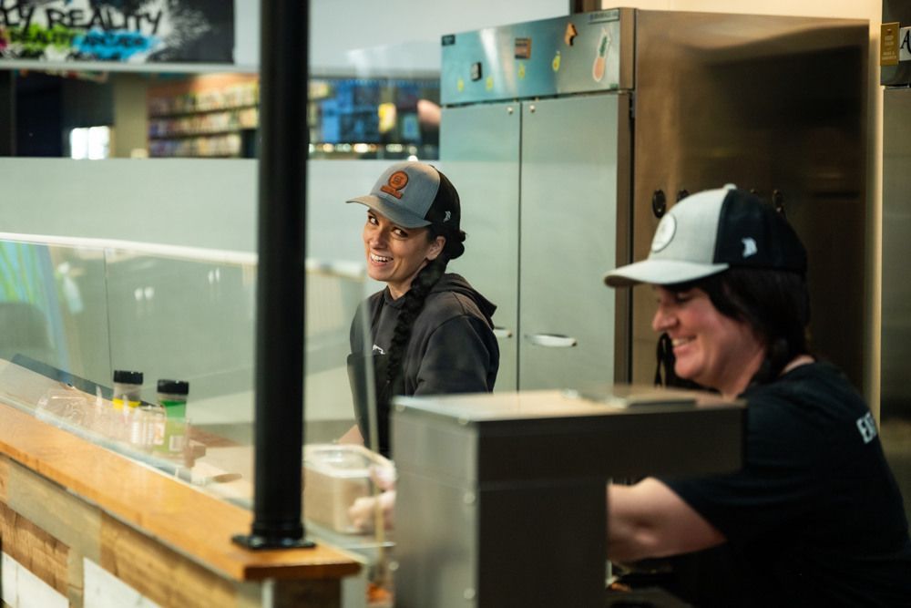 Two people in hats smile behind a counter. One wears a black hat with an orange logo.
