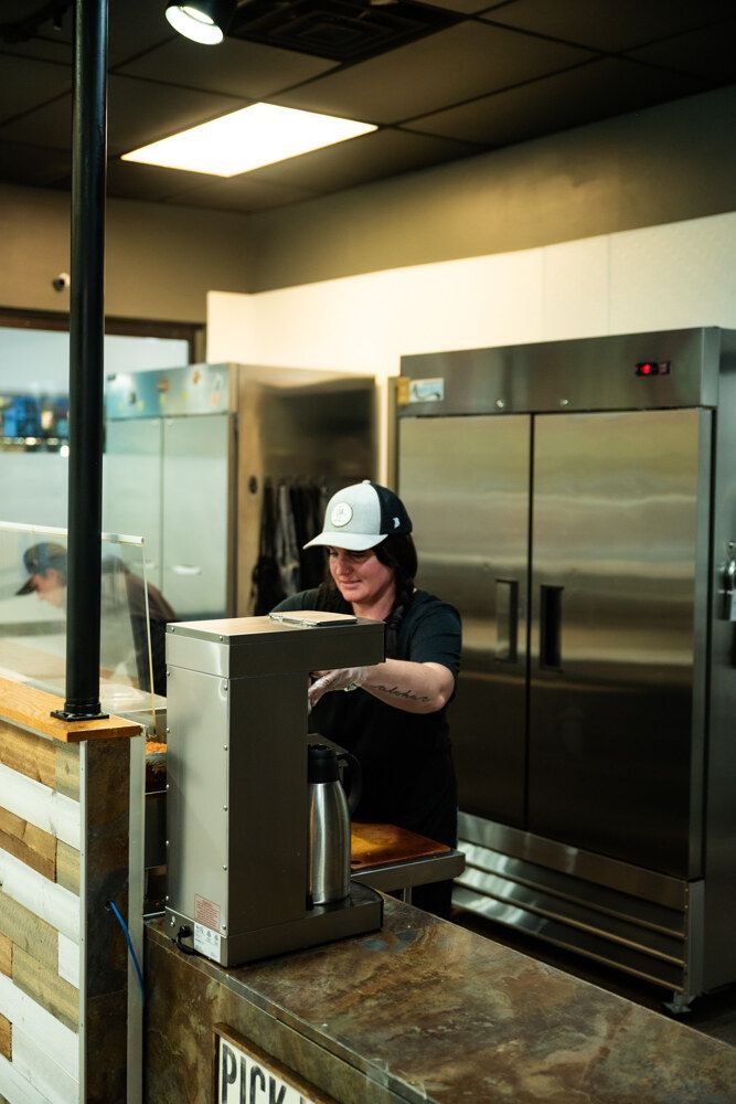 Person in a baseball cap pouring from a coffee machine at a counter in a commercial kitchen.